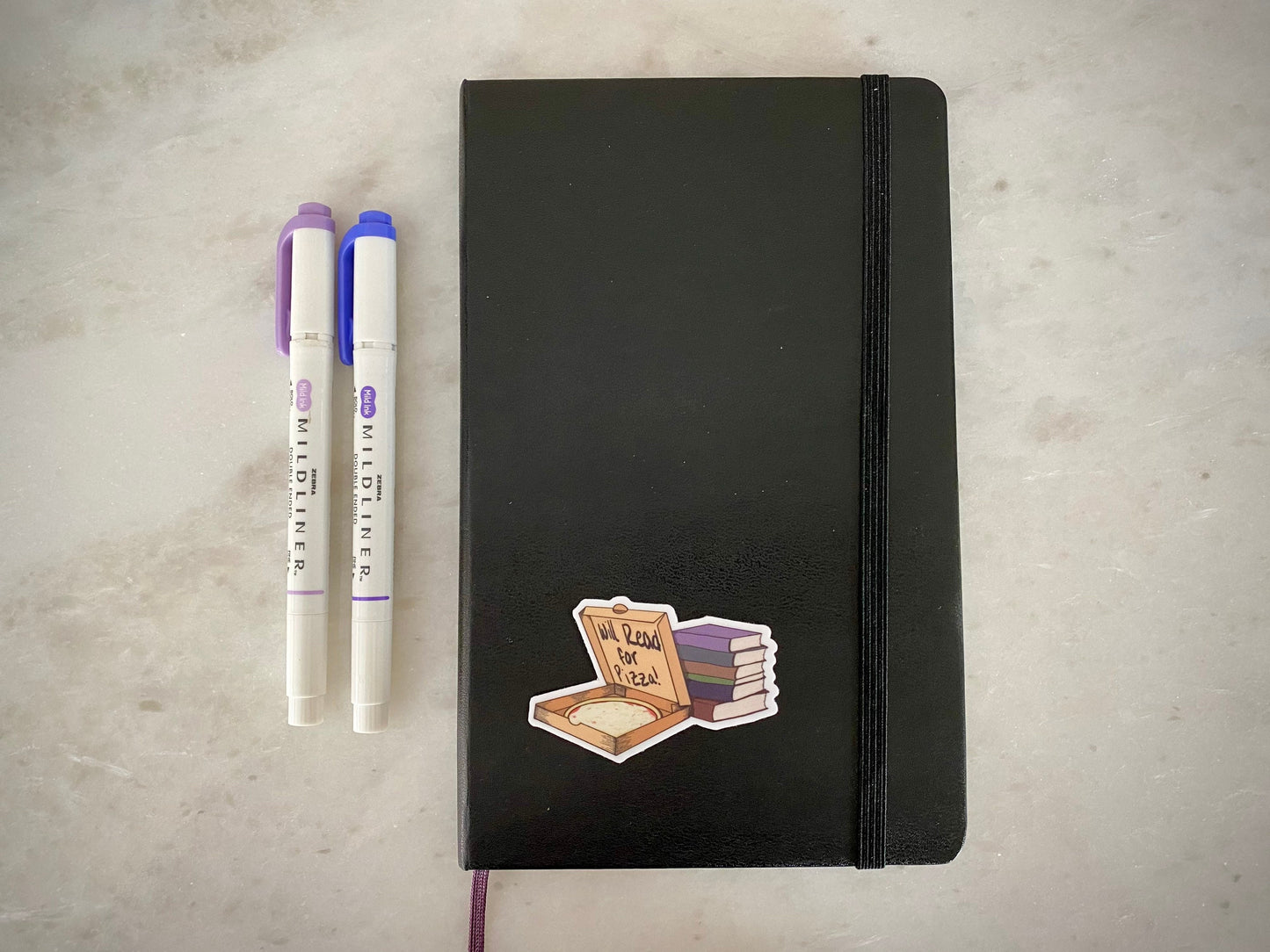 hand holding a sticker showing a pizza box with a stack of books with text "will read for pizza" on a black notebook with purple and blue markers sitting on a marble surface