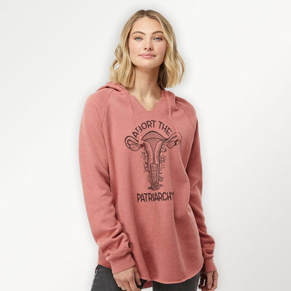 blond woman wearing a dusty rose colored hooded sweatshirt with a black screen printed floral uterus graphic and "abort the patriarchy" text
