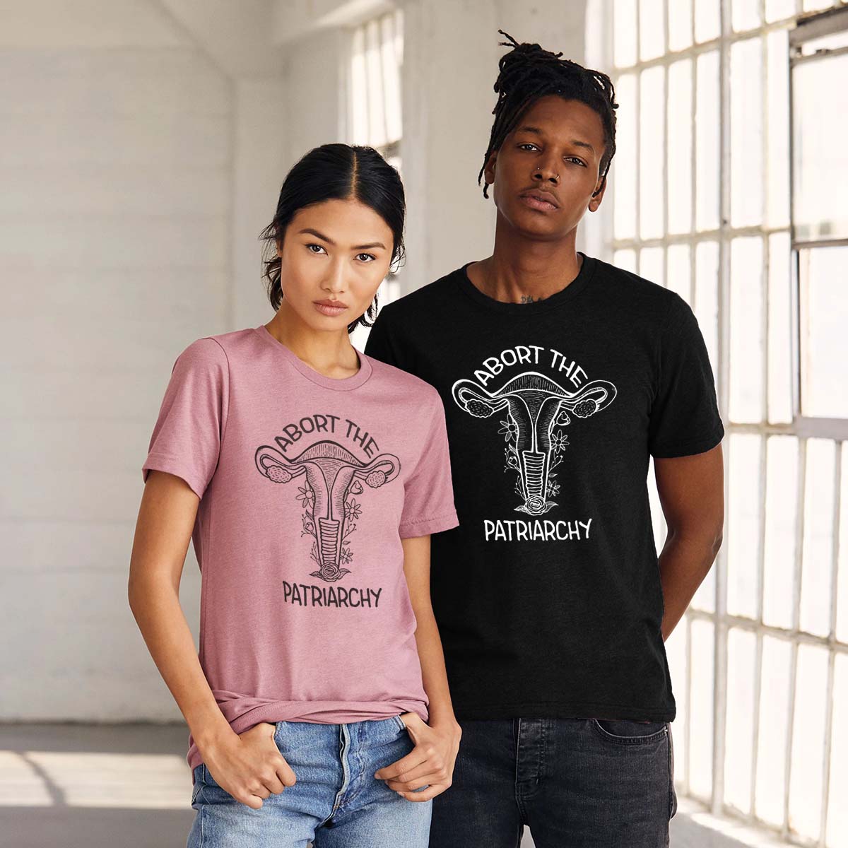 an asian woman wearing an orchid colored t-shirt with a white screen printed floral uterus graphic and "abort the patriarchy" text  standing next to a black man wearing a black t-shirt with a white screen printed floral uterus graphic and "abort the patriarchy" text