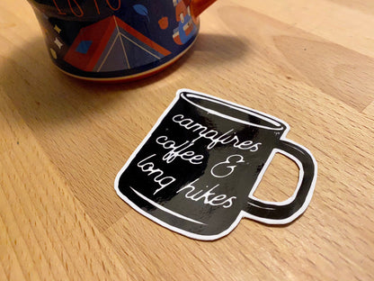 sticker of a black camp coffee mug with the text "campfires, coffee, and long hikes" on light wood background