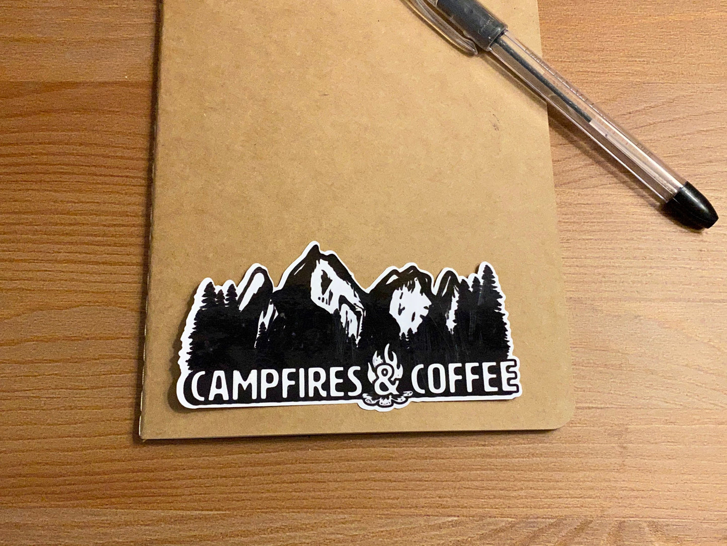 black and white vinyl sticker with a mountain and tree illustration and the text "campfires and coffee" sitting on a brown background